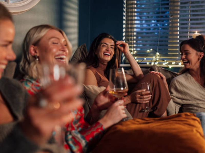 A group of women sit on a couch having girls night in, holding wine and laughing.